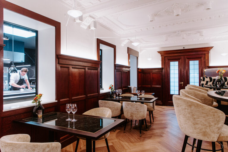 Sfeervol restaurantinterieur met donkerhouten lambrisering, marmeren tafels en een chef in een open keuken.