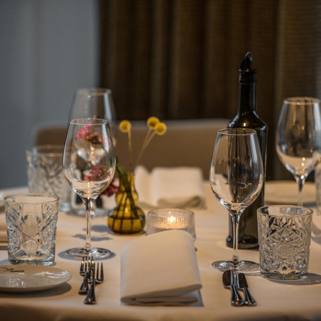 Gedekte restauranttafel met wijnglazen, kristallen glazen, bestek, witte servetten, kaars, bloemen en een fles wijn, op het bordje COMPAS.