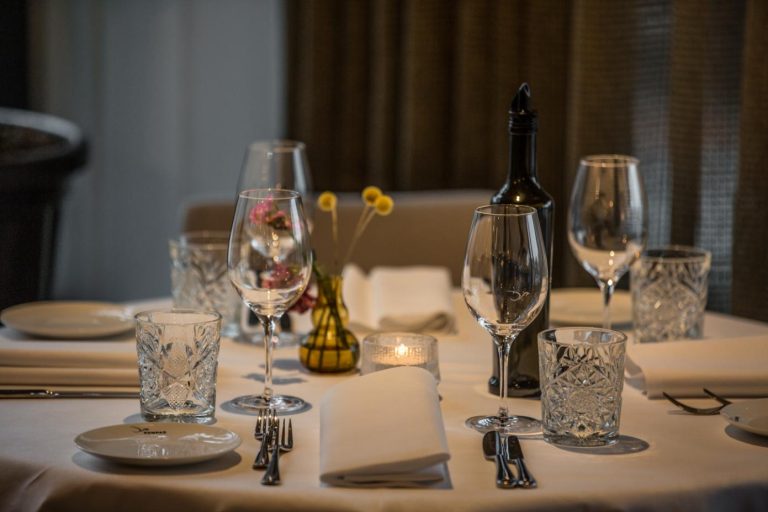 Gedekte restauranttafel met wijnglazen, kristallen glazen, bestek, witte servetten, kaars, bloemen en een fles wijn, op het bordje COMPAS.