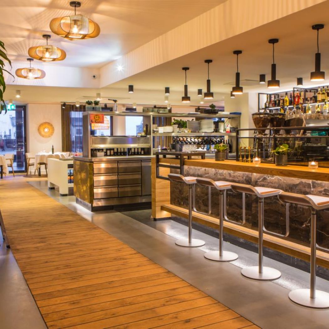 Modern restaurant- en barinterieur met lange verlichte bar, strakke barkrukken en gedekte tafels.