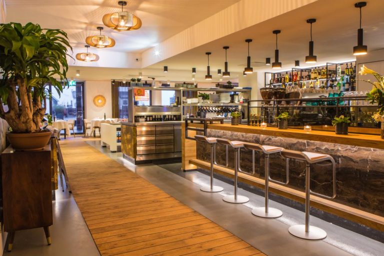 Modern restaurant- en barinterieur met lange verlichte bar, strakke barkrukken en gedekte tafels.