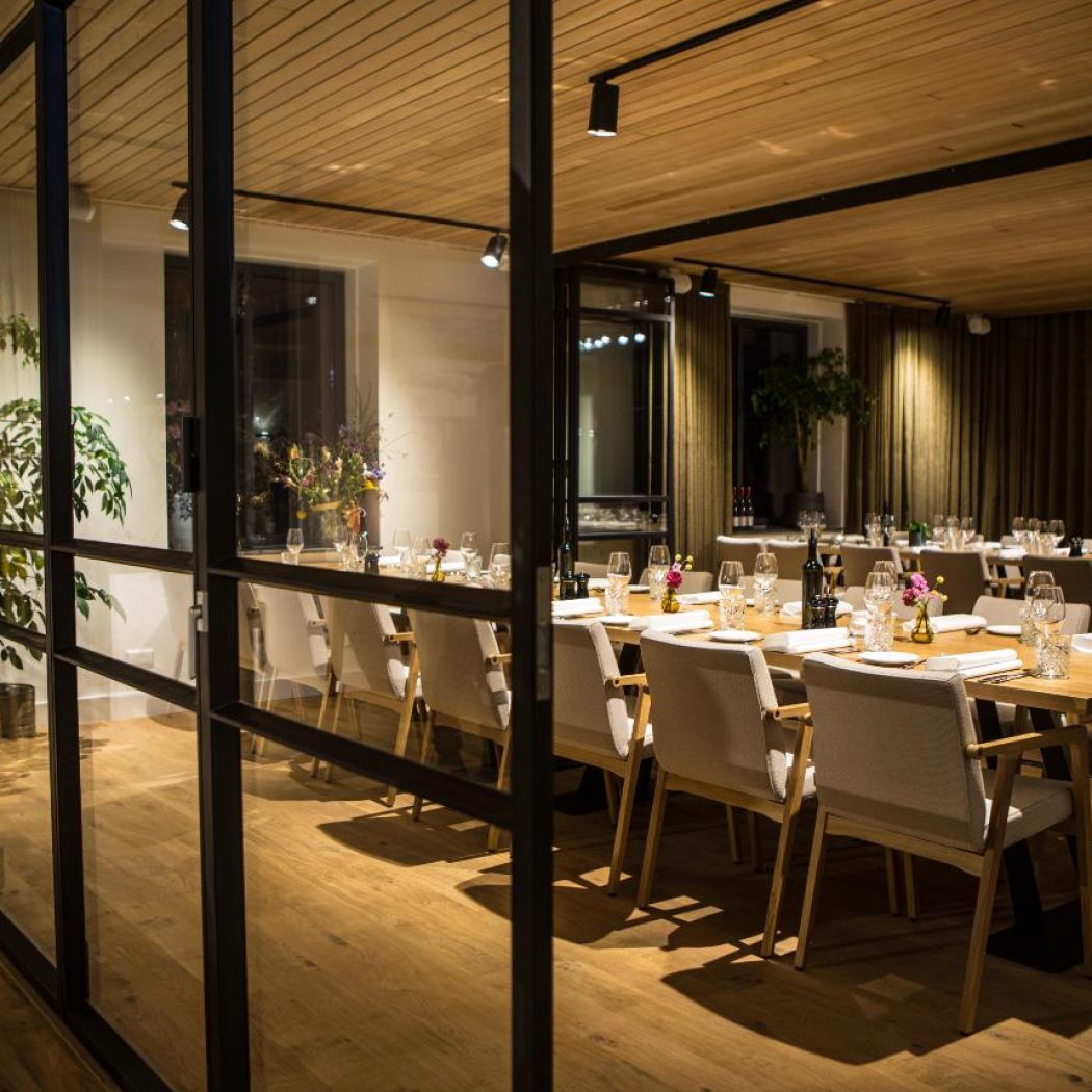 Sfeervolle, modern ingerichte dinerzaal met houten tafels, beige stoelen en een glazen scheidingswand.