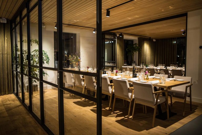 Sfeervolle, modern ingerichte dinerzaal met houten tafels, beige stoelen en een glazen scheidingswand.