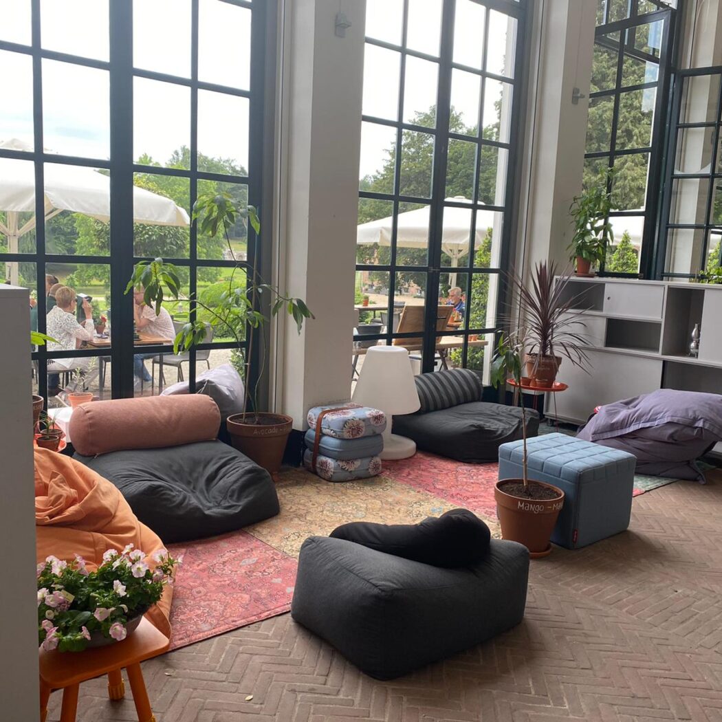 Ontspannen loungehoek met zitzakken en planten, met uitzicht op een tuin en gasten onder parasols.