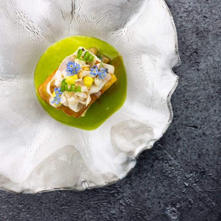 Verfijnd gerecht: toast met fijngesneden topping, blauwe vergeet-mij-nietjes, gele stippen en groene saus op een wit golvend bord.
