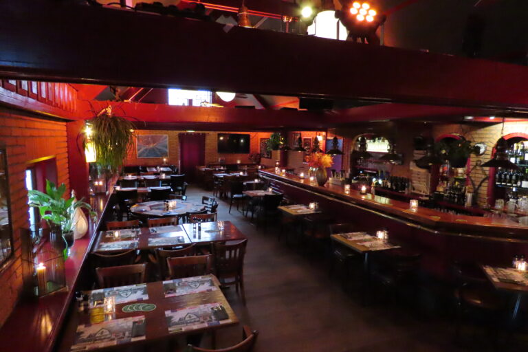 Warm verlicht restaurant interieur met rode bakstenen muren, eettafels en een lange bar met kaarsen.