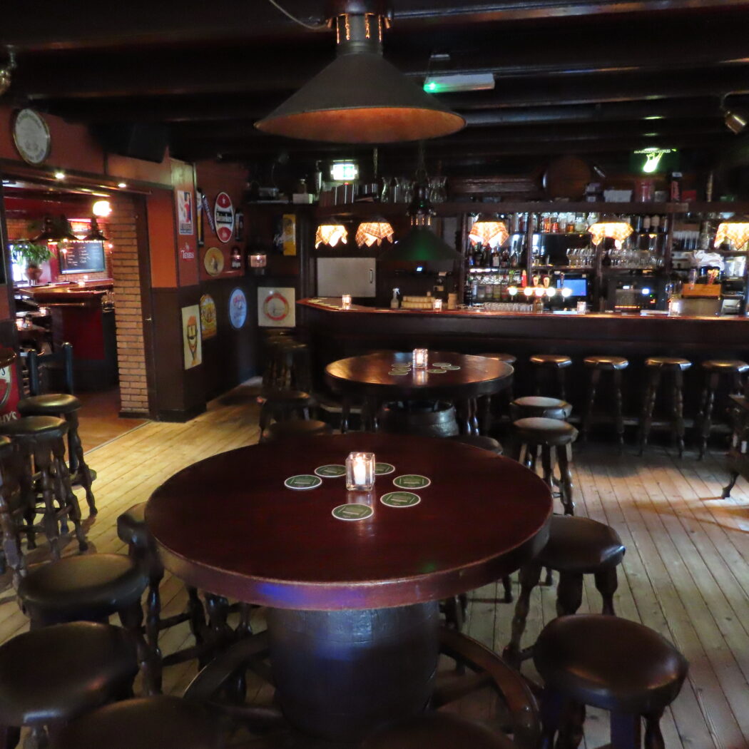 Warm verlicht pubinterieur met houten bar, barkrukken en ronde tafels met kaarslicht.