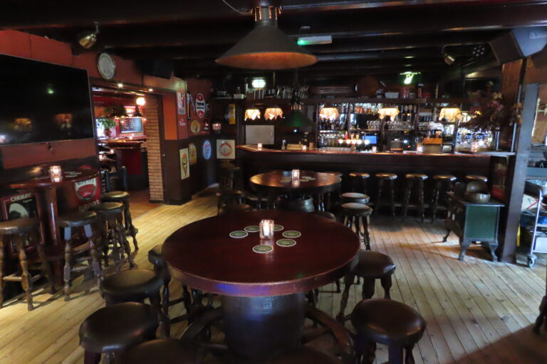 Warm verlicht pubinterieur met houten bar, barkrukken en ronde tafels met kaarslicht.