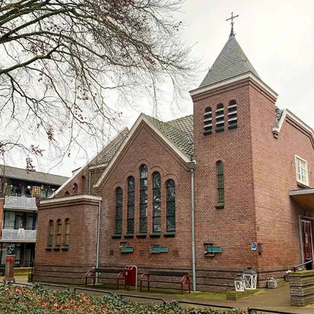 Rode bakstenen kerk met een spitse toren, kruis, boogramen en geparkeerde fietsen onder kale bomen.