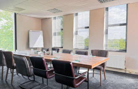 Ruime, lichte vergaderzaal met robuuste houten tafel, diverse stoelen, grote ramen en een whiteboard.