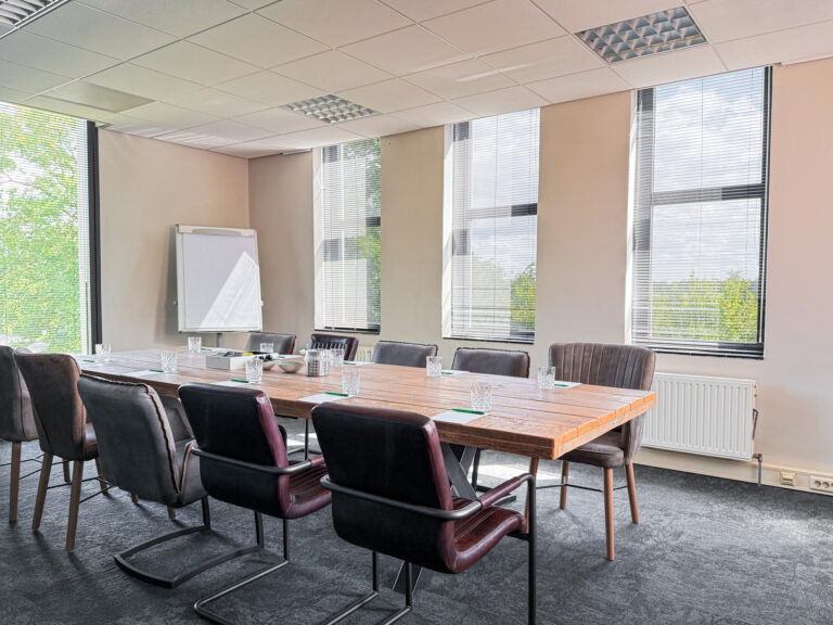 Ruime, lichte vergaderzaal met robuuste houten tafel, diverse stoelen, grote ramen en een whiteboard.