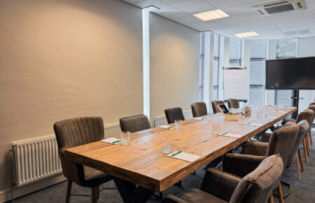 Een moderne vergaderruimte met lange houten tafel, gestoffeerde stoelen, een flipchart en groot tv-scherm.