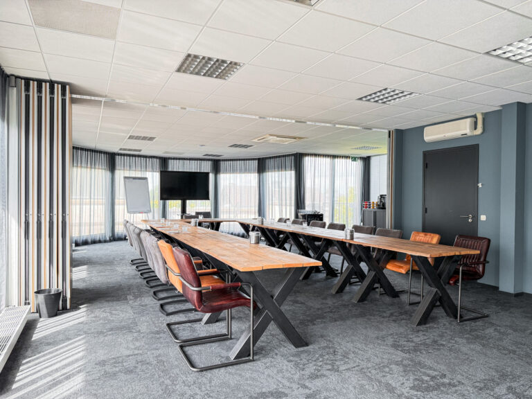 Stijlvolle vergaderruimte met lange houten tafels op metalen X-poten, diverse stoelen en groot scherm.