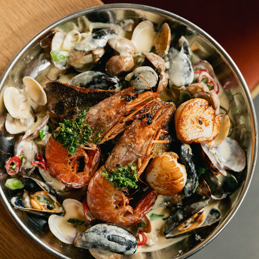 Zilveren schaal met gebakken garnalen, coquilles, mosselen en kokkels in romige saus, gegarneerd met groene kruiden op een houten tafel.
