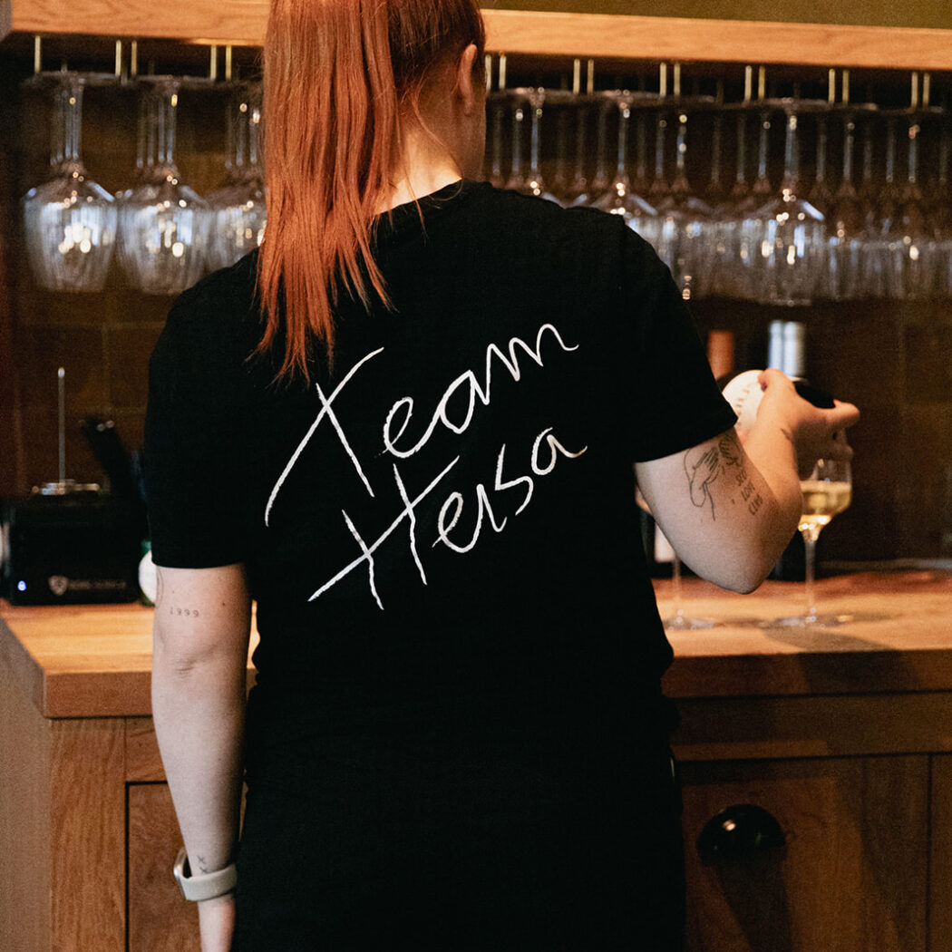 Roodharig teamlid met Team Hesa shirt staat bij een houten bar met wijnglazen.