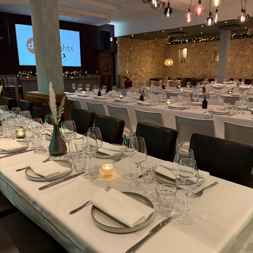 Elegante restaurantzaal met wit gedekte tafels, glazen, kaarslicht en twee schermen met d hts.