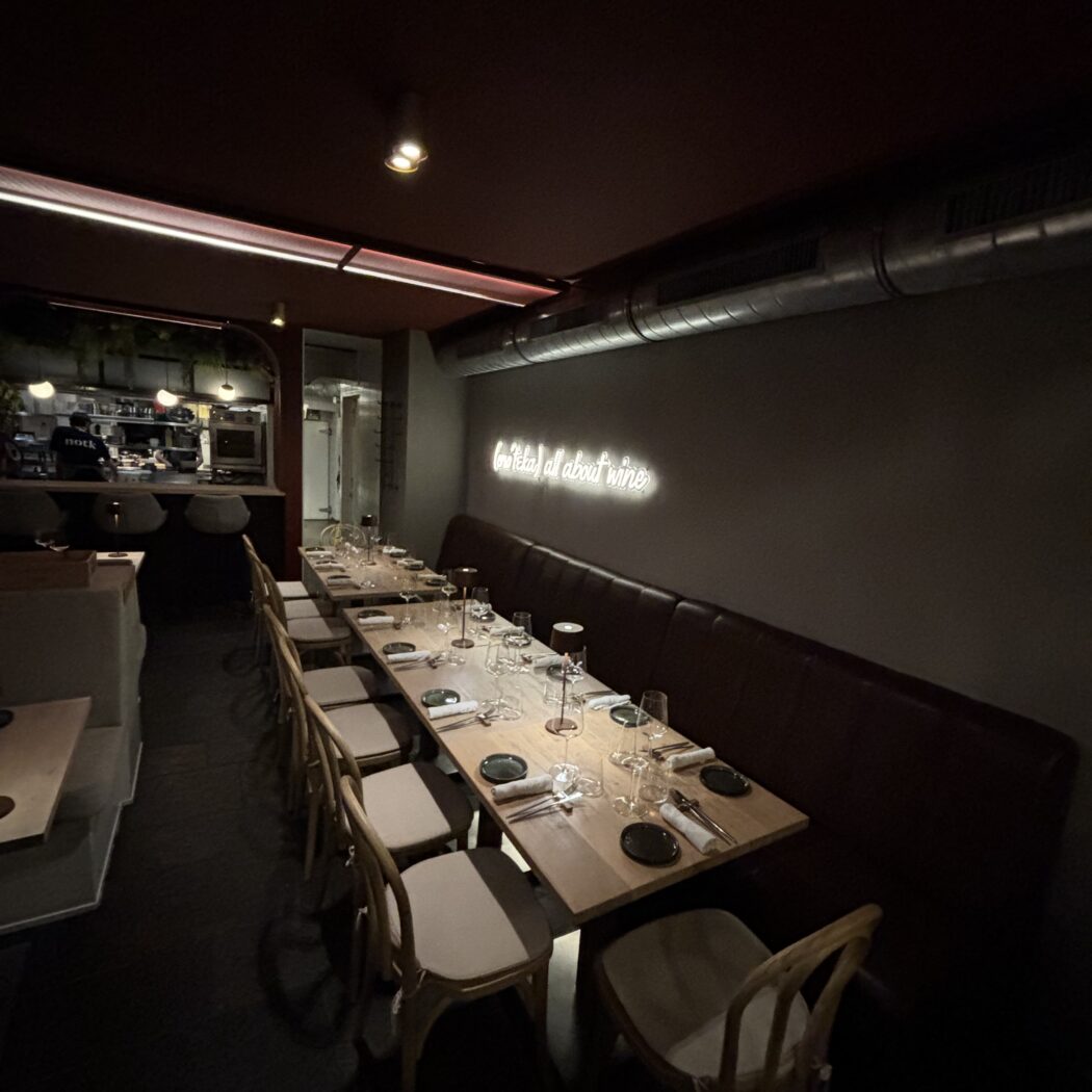 Sfeervol restaurant met lange gedekte houten tafel, houten stoelen en neontekst Its all about wine.