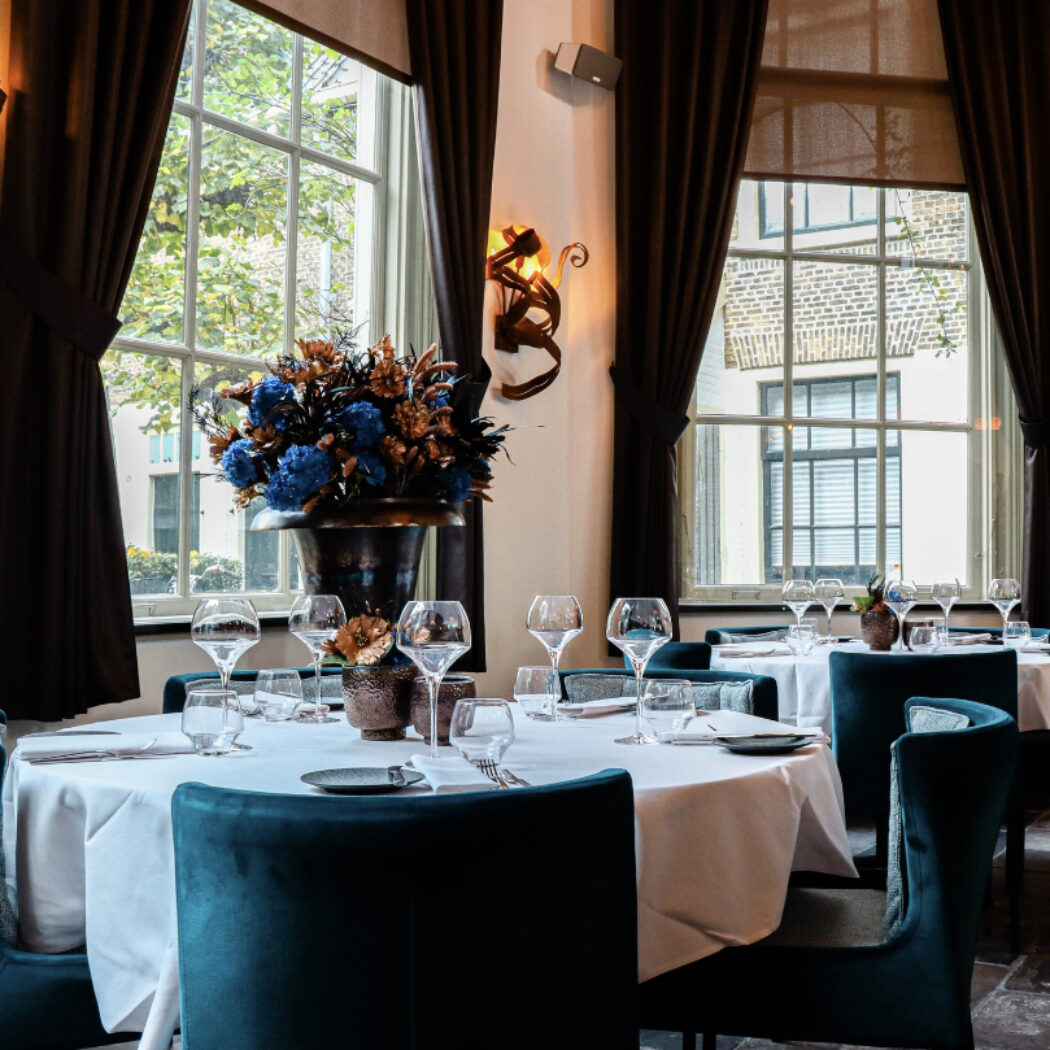 Elegant restaurantinterieur met gedekte tafels, blauwe fluwelen stoelen en een bloemstuk met blauwe en bronzen bloemen.