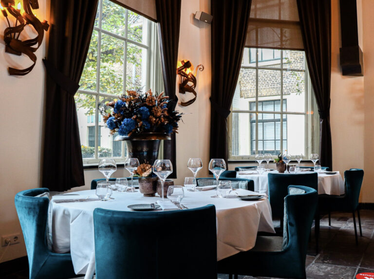 Elegant restaurantinterieur met gedekte tafels, blauwe fluwelen stoelen en een bloemstuk met blauwe en bronzen bloemen.