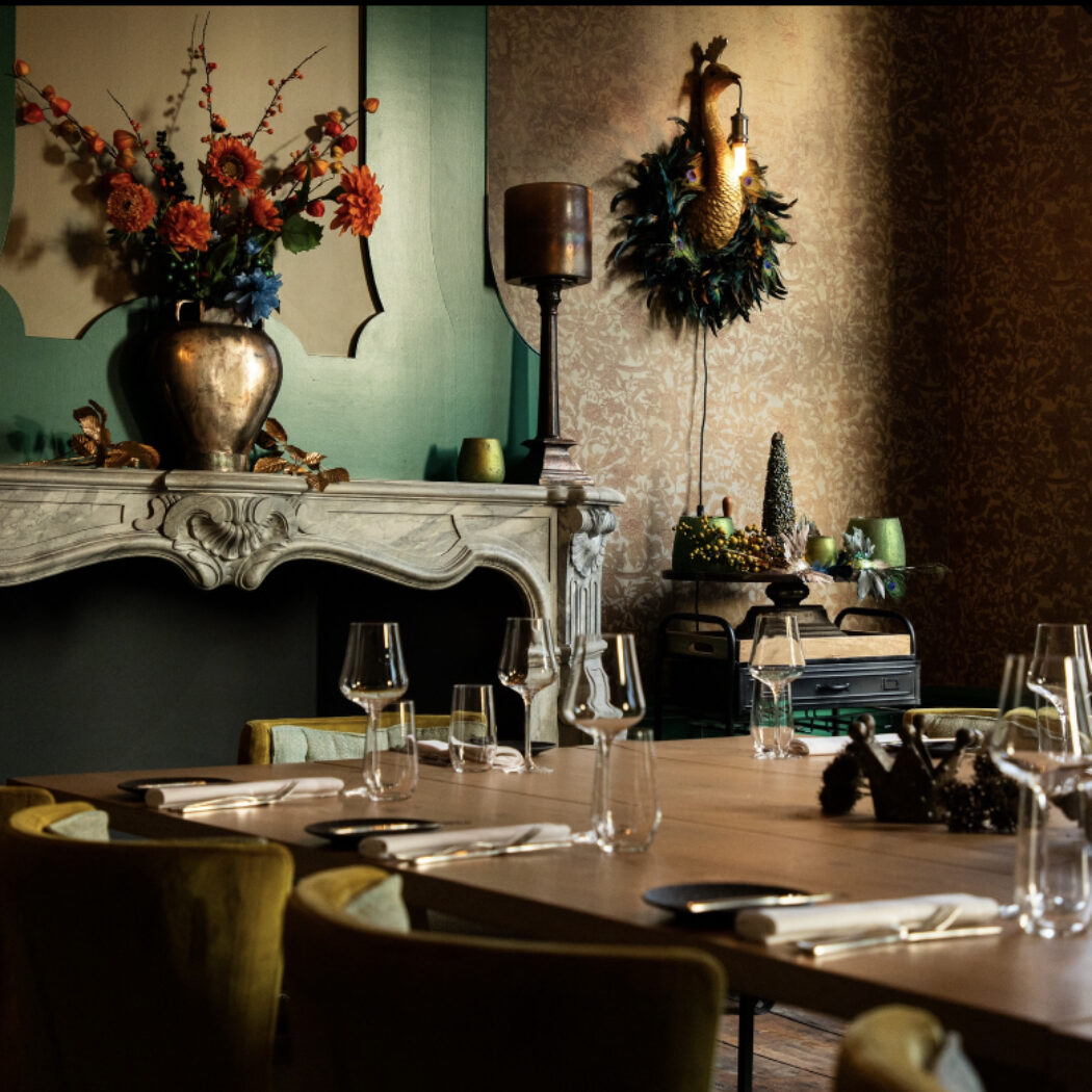 Sfeervolle, luxe eetkamer met gedekte lange houten tafel, glazen, mosterdgele stoelen, open haard en kleurrijke bloemen.