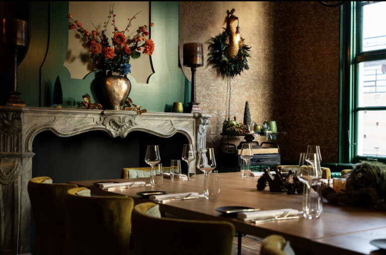Sfeervolle, luxe eetkamer met gedekte lange houten tafel, glazen, mosterdgele stoelen, open haard en kleurrijke bloemen.