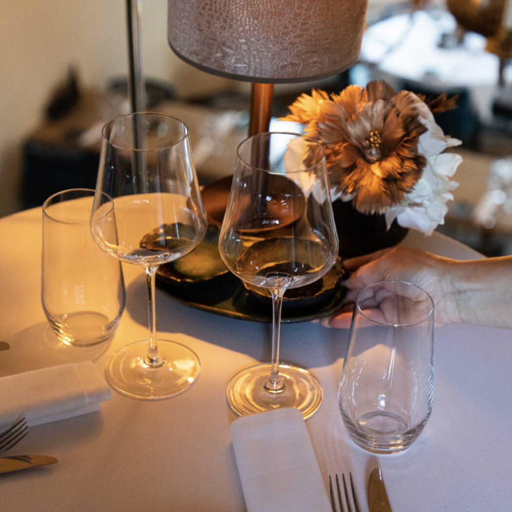 Gedekte restauranttafel met wijnglazen en bestek, waarbij een hand een bloemstuk schikt.