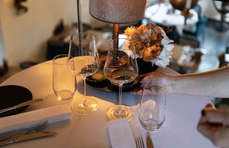 Gedekte restauranttafel met wijnglazen en bestek, waarbij een hand een bloemstuk schikt.