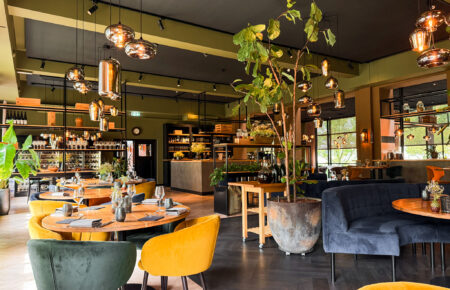 Modern restaurantinterieur met ronde houten tafels, gele en donkergroene velours stoelen, en sfeervolle hanglampen.