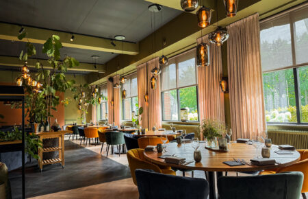 Sfeervol restaurantinterieur met ronde houten tafels, gekleurde stoelen, warme hanglampen en uitzicht op groen.