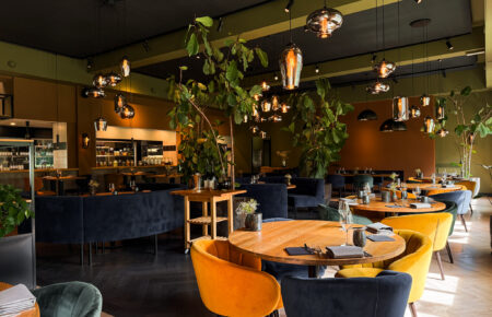 Sfeervol restaurant met ronde houten tafels, donkerblauwe en okergele velvet stoelen, en diverse glazen hanglampen.