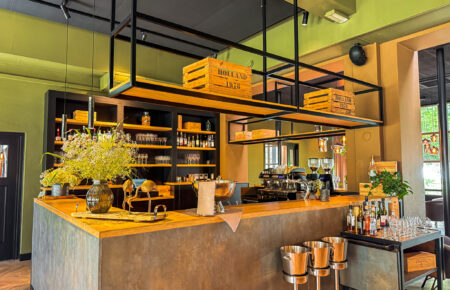 Interieur van een moderne bar met houten blad, espressoapparaat, planken vol flessen en ‘HOLLAND 1978’ kratten.