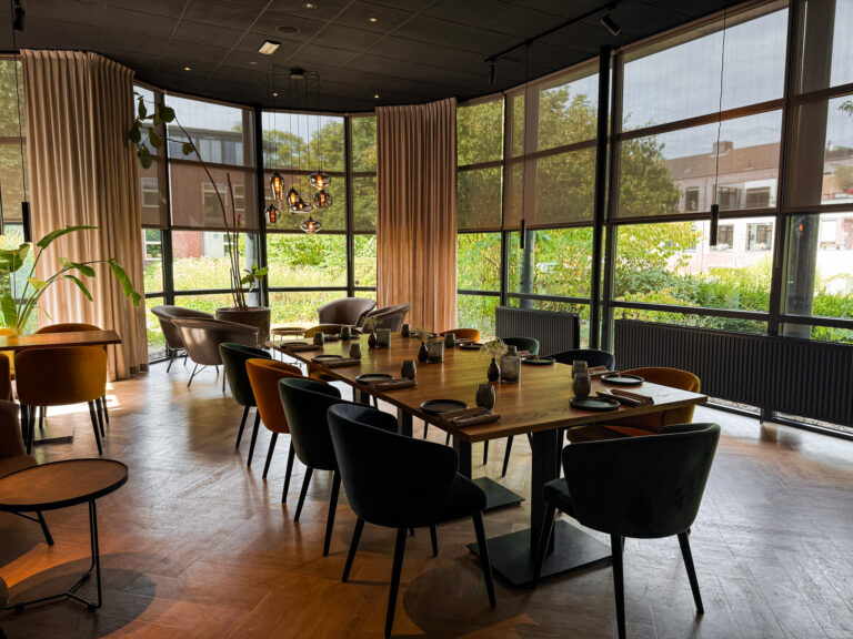 Moderne restaurantruimte met lange houten tafel voor 12, diverse fluwelen stoelen, ramen met zonwering en beige gordijnen.