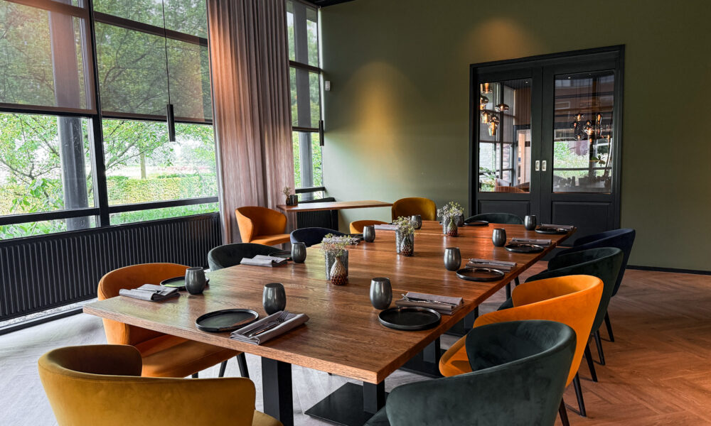 Moderne vergaderruimte met lange houten tafel, gedekte plaatsen, oranje en donkergroene velvet stoelen en grote ramen.