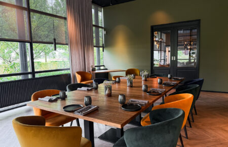 Moderne vergaderruimte met lange houten tafel, gedekte plaatsen, oranje en donkergroene velvet stoelen en grote ramen.