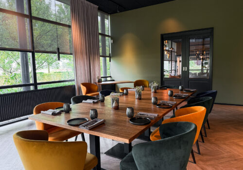 Moderne vergaderruimte met lange houten tafel, gedekte plaatsen, oranje en donkergroene velvet stoelen en grote ramen.
