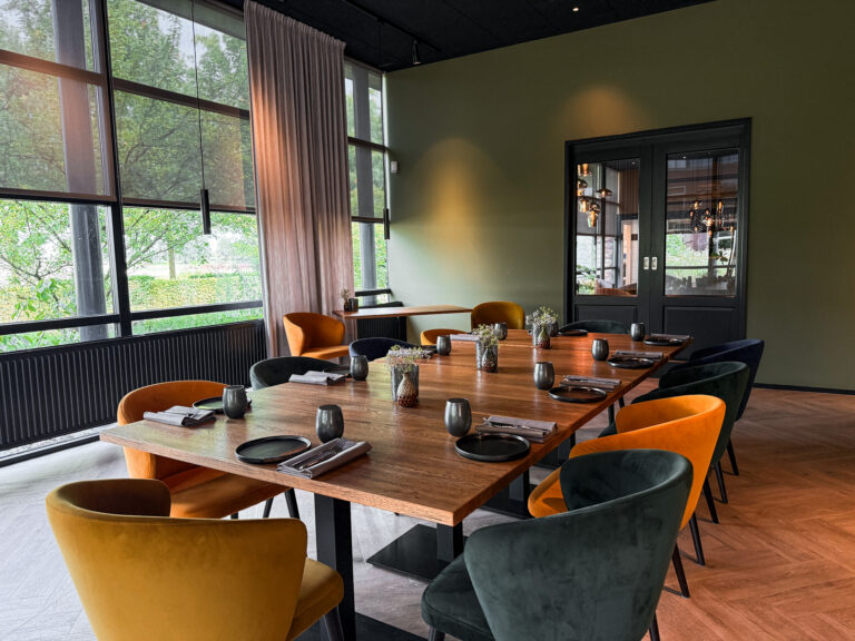Moderne vergaderruimte met lange houten tafel, gedekte plaatsen, oranje en donkergroene velvet stoelen en grote ramen.