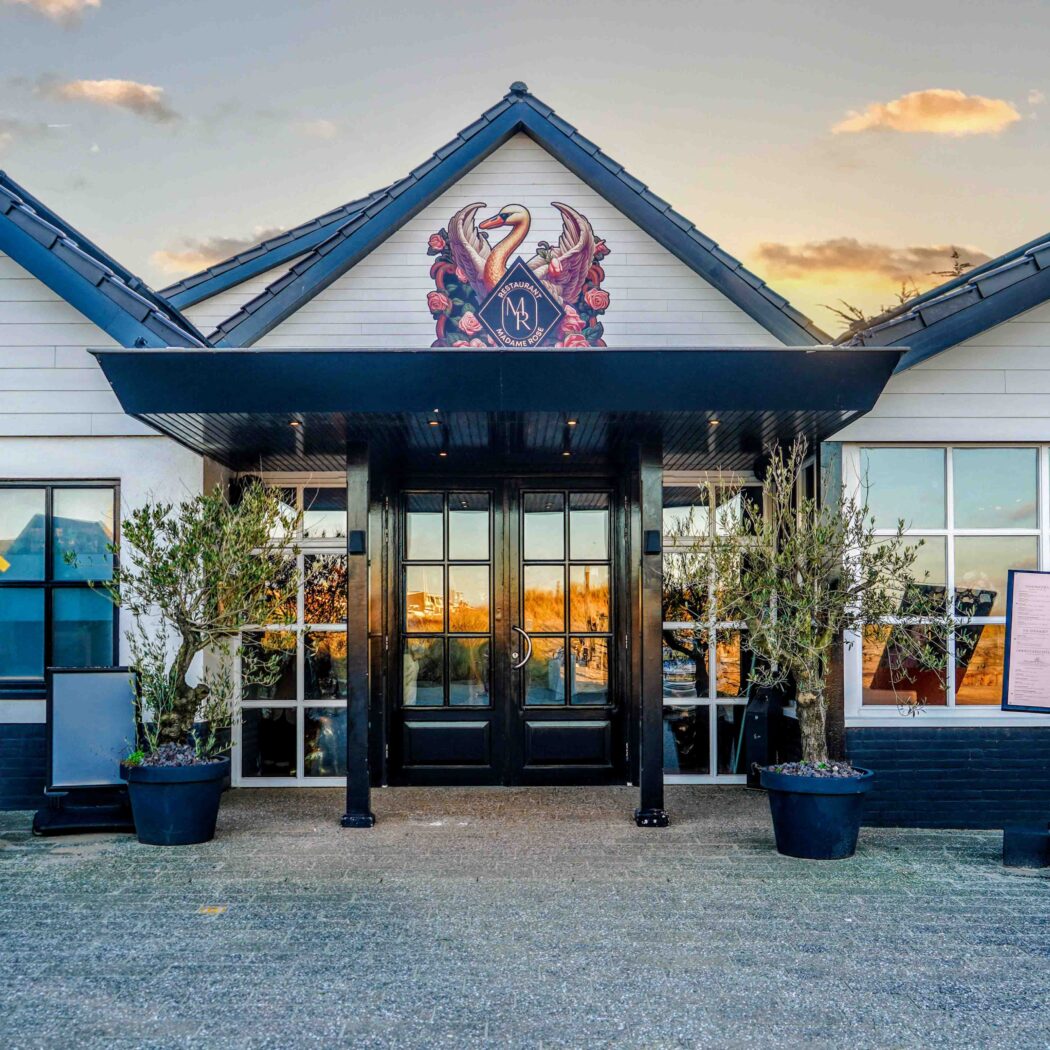 Ingang van Restaurant Madame Rosie met dubbele zwarte deuren, olijfbomen en het bord ‘Everyday is a day of wine and roses’.