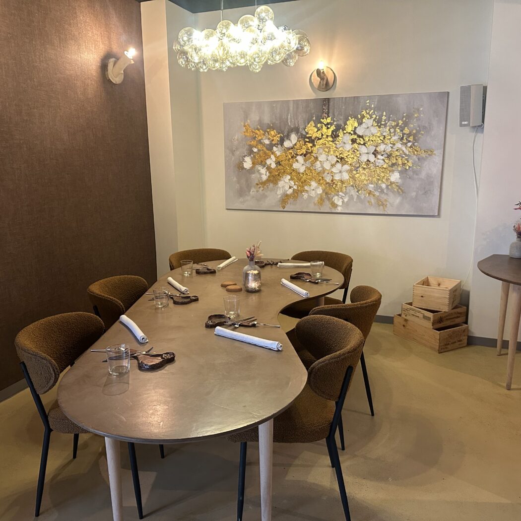 Ovale gedekte eettafel met bruine stoelen, grote gouden wandkunst en glazen kroonluchter in een restaurant.