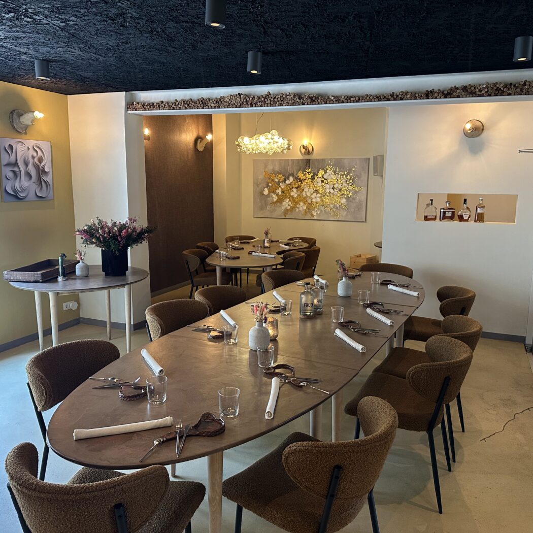 Sfeervol restaurantinterieur met lange ovale tafel, bruine bouclé stoelen, bloemen en moderne kunst aan de olijfgroene muren.