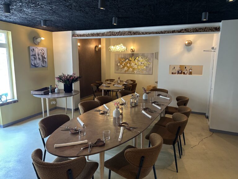 Sfeervol restaurantinterieur met lange ovale tafel, bruine bouclé stoelen, bloemen en moderne kunst aan de olijfgroene muren.
