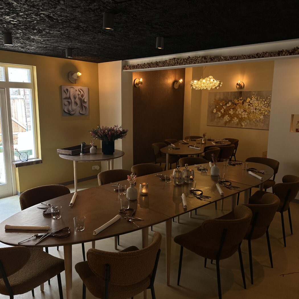Sfeervolle restaurantzaal met lange gedekte houten tafel, bruine stoelen, gouden accenten en muurkunst.