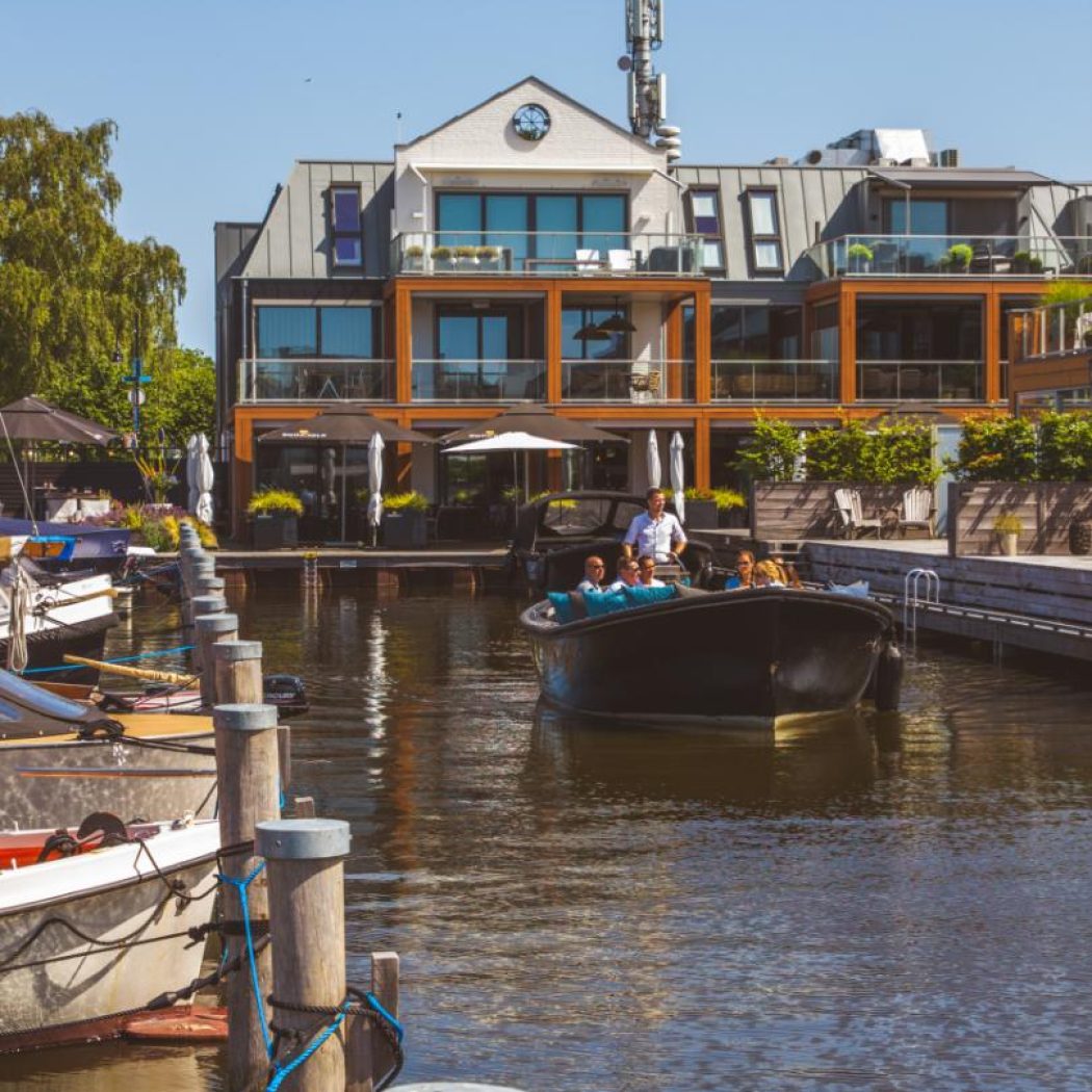 Een donkere sloep met passagiers vaart op een zonnige dag langs een modern pand met terrassen aan het water.