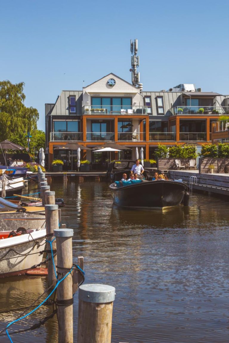 Een donkere sloep met passagiers vaart op een zonnige dag langs een modern pand met terrassen aan het water.