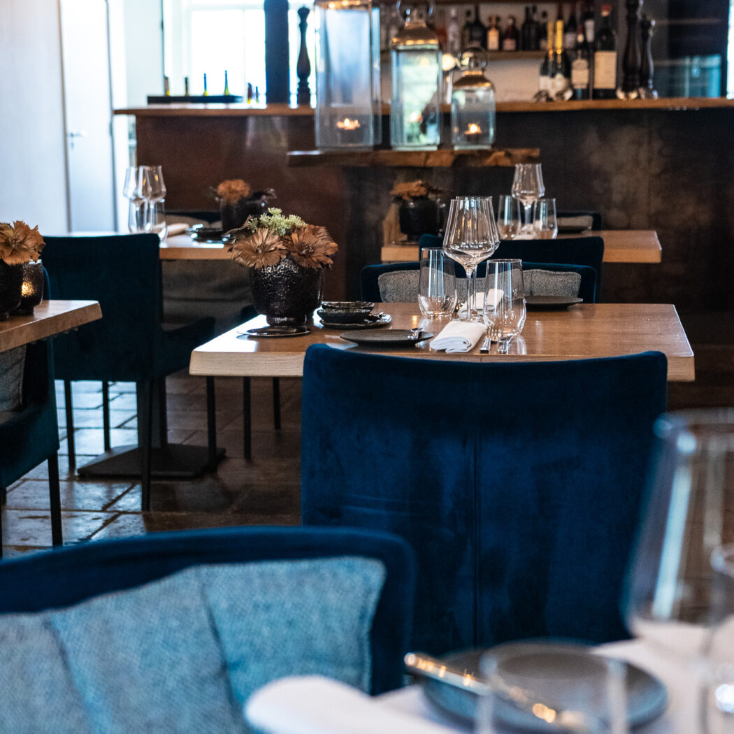 Sfeervol restaurantinterieur met gedekte houten tafels, donkerblauwe velvet stoelen en een verlichte bar.