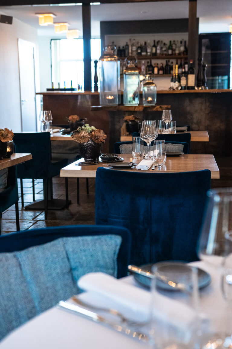 Sfeervol restaurantinterieur met gedekte houten tafels, donkerblauwe velvet stoelen en een verlichte bar.