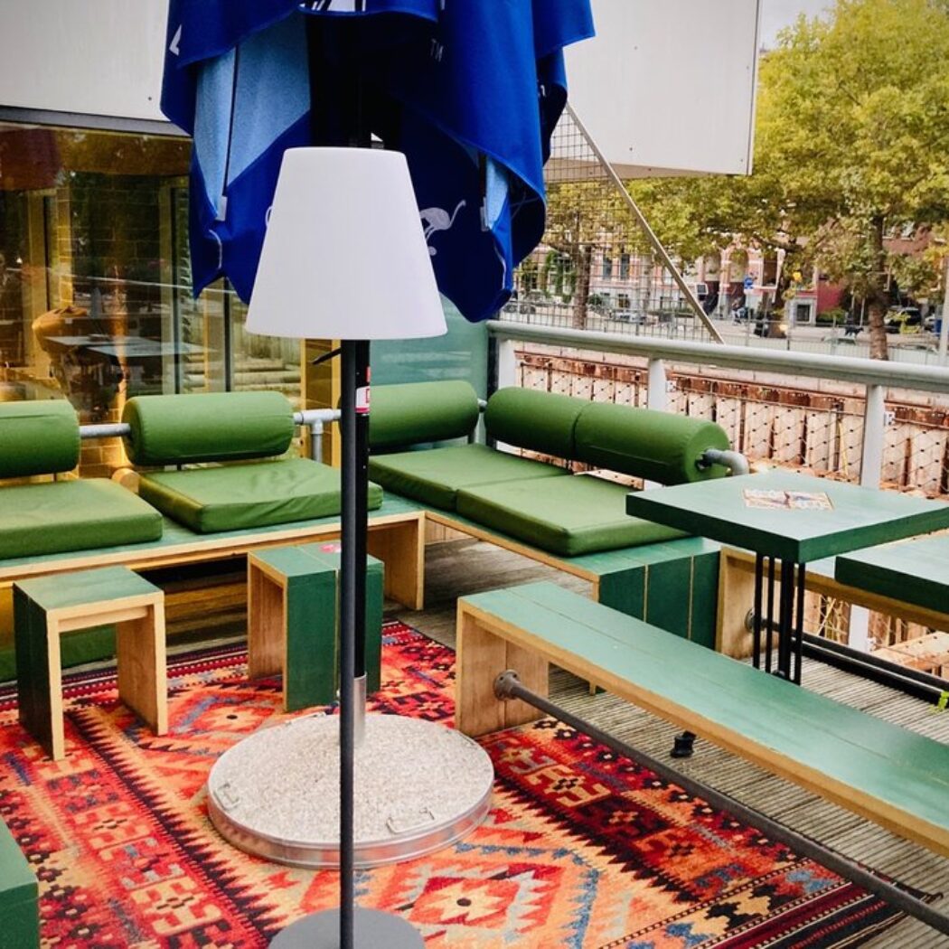 Buitenterras met groene zitbanken, houten tafels en een staande lamp op een kleurrijk rood-blauw tapijt, bij een kanaal.