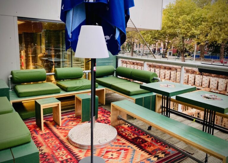 Buitenterras met groene zitbanken, houten tafels en een staande lamp op een kleurrijk rood-blauw tapijt, bij een kanaal.
