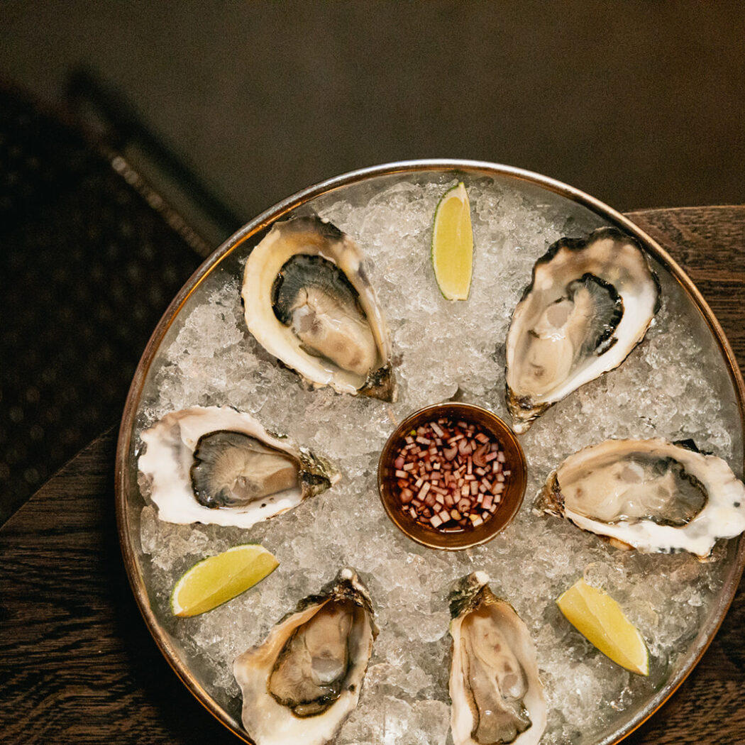 Rond dienblad met zes verse oesters op crushed ijs, geserveerd met limoenpartjes en rode ui mignonette op een houten tafel.