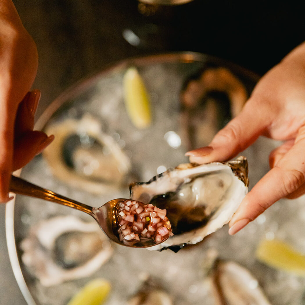 Handen voegen rode uienmignonette toe aan een open oester op een ijsschotel met limoenpartjes.