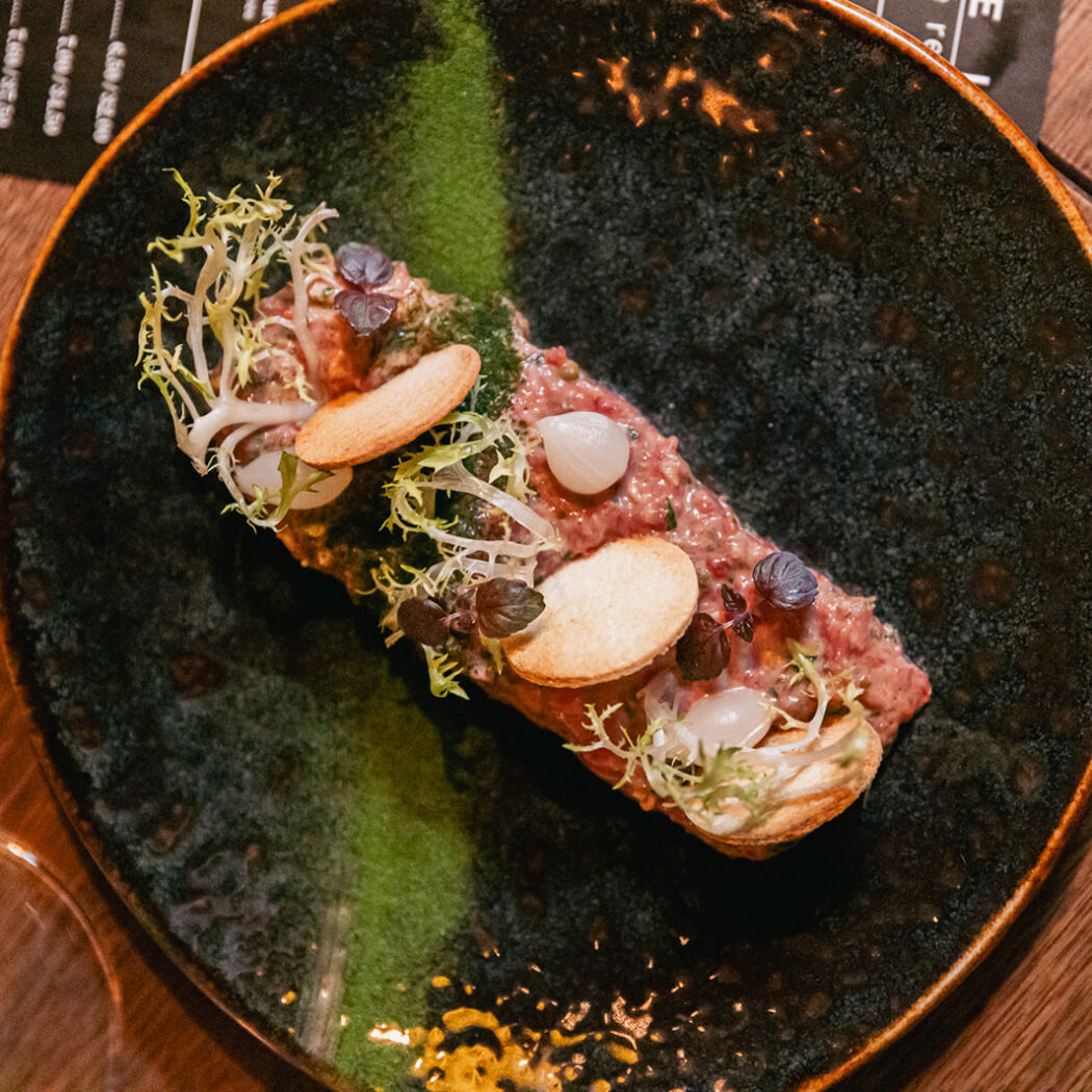 Steaktartaar met frisée, kleine uitjes en knapperige toast op een donker gespikkeld bord met groene streep.